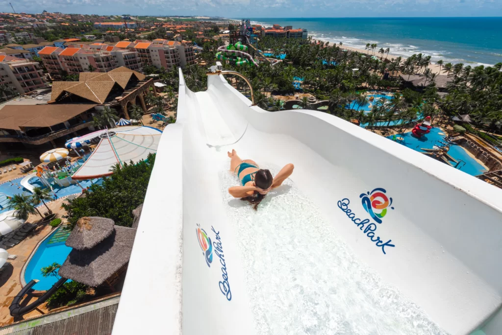 a imagem mostra uma mulher descendo no Insano no Aqua Park do Beach Park nas férias de julho. Ao fundo da imagem, há a paisagem da Praia do Porto das Dunas, no Ceará