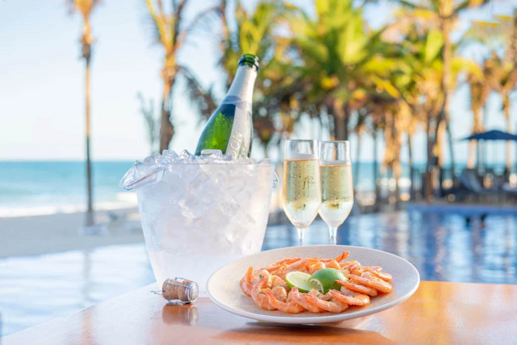 a imagem mostra um prato de camarão com taças de champagne e um um balde de gelo à beira-mar com coqueiros ao fundo da imagem na praia do Beach Park