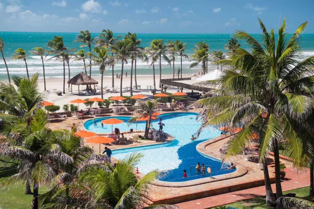 a imagem mostra parte da estrutura do Oceani Beach Park Resort com piscina, pé na areia e mar da Praia do Porto das Dunas com coqueiros ao fundo