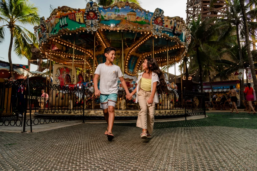 a imagem mostra duas crianças sorrindo de mãos dadas e caminhando em frente ao carrossel da Vila Azul do Mar, do Beach Park, no Ceará