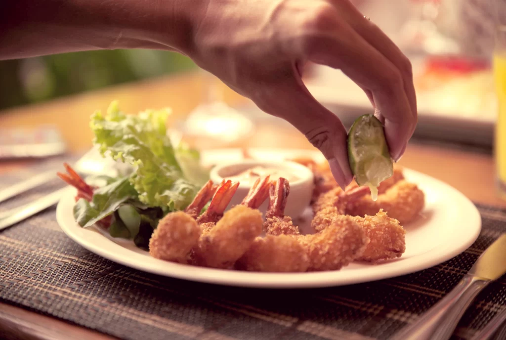 a imagem mostra um prato de culinária regional com camarões, salada, e a mão de uma pessoa espremendo limão por cima