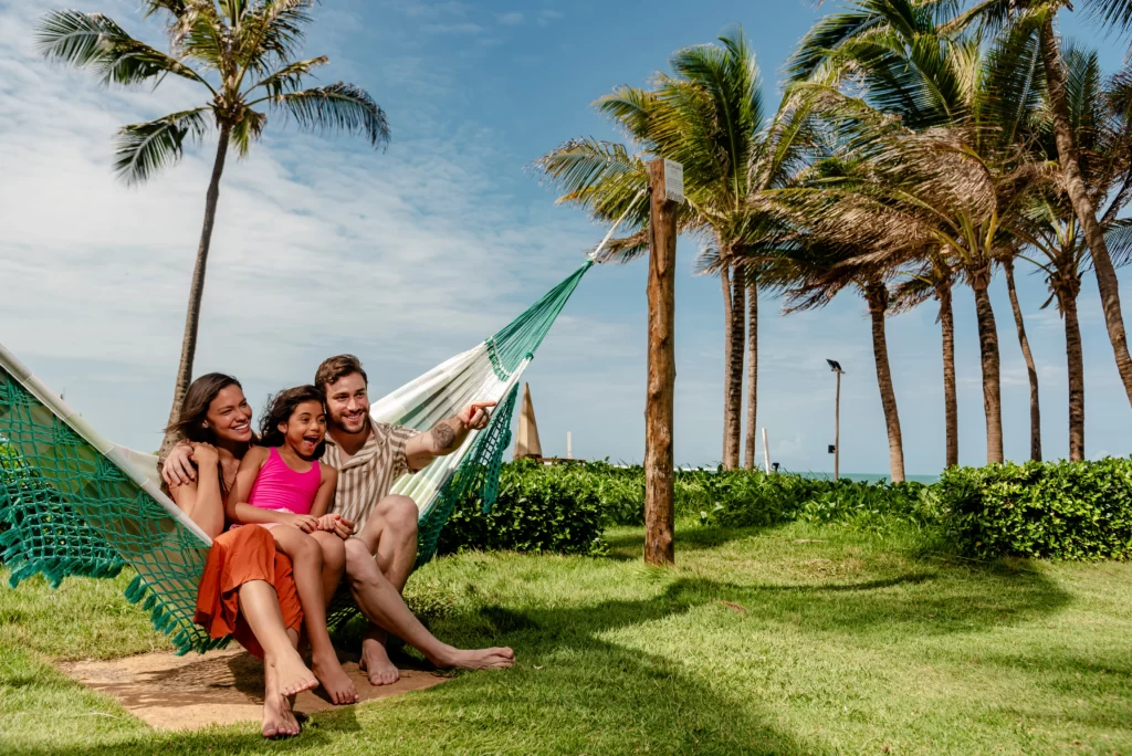 a imagem mostra uma família se divertindo no balanço de uma rede ao ar livre com coqueiros, gramado e vista para o mar no litoral do ceará